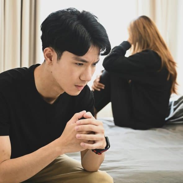 A man sitting on a bed with clasped hands while a woman sits in the background.