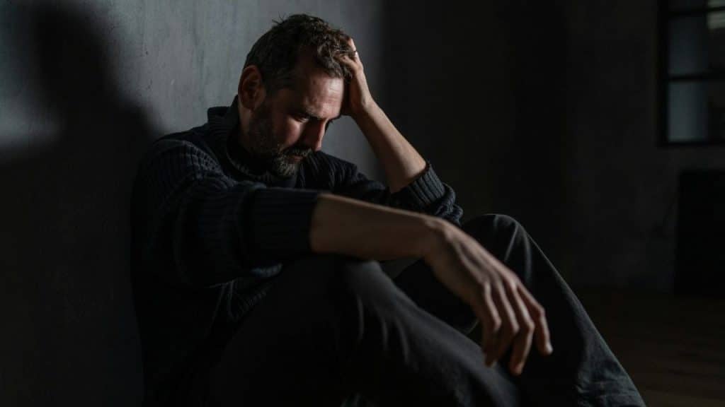 Man sitting on floor, head in hand, in a dark, dimly lit room.