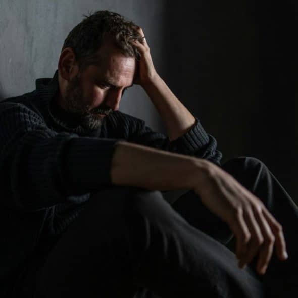 Man sitting on floor, head in hand, in a dark, dimly lit room.