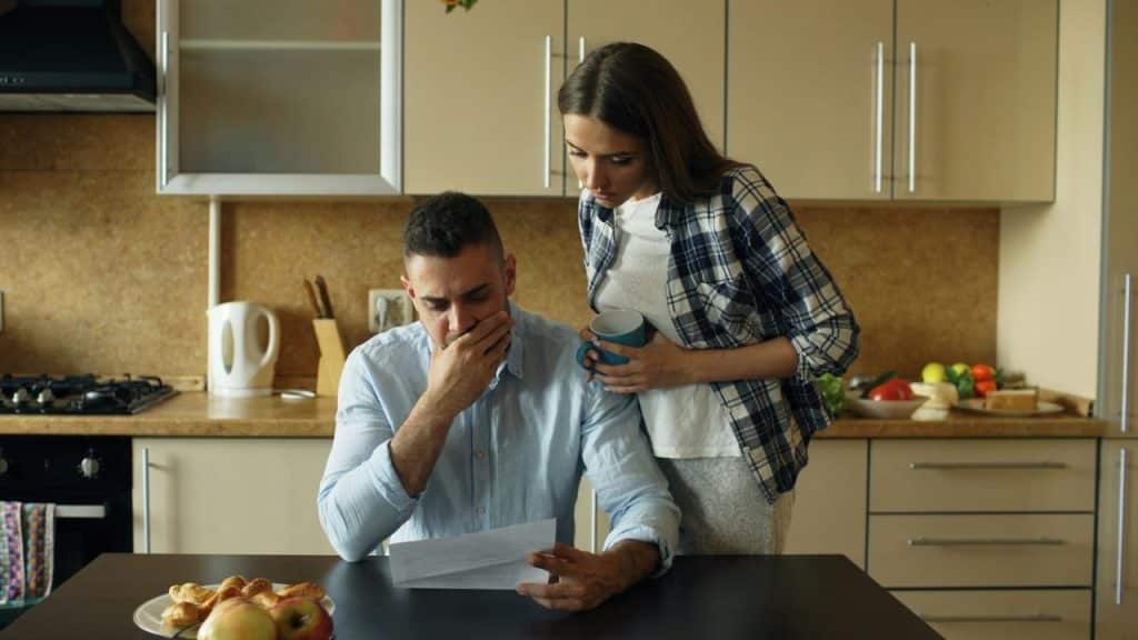 A distressed man reading a paper at a kitchen table with a woman looking over his shoulder.