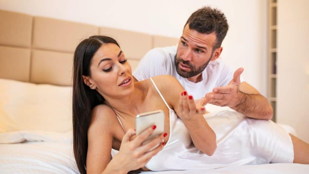 A man arguing with a woman on a bed who is holding and looking at a phone.