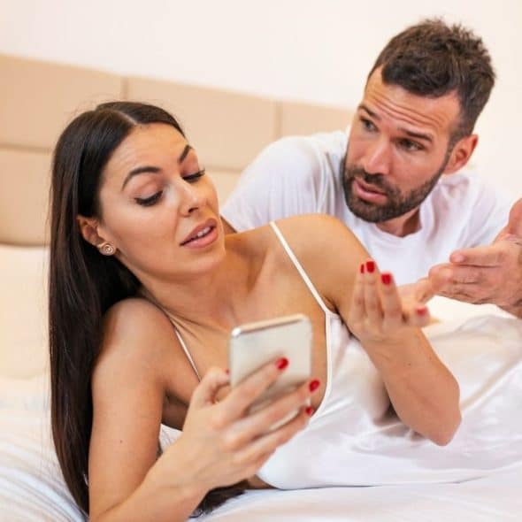 Woman in satin slip looking at phone while man argues/gestures behind her in bed.