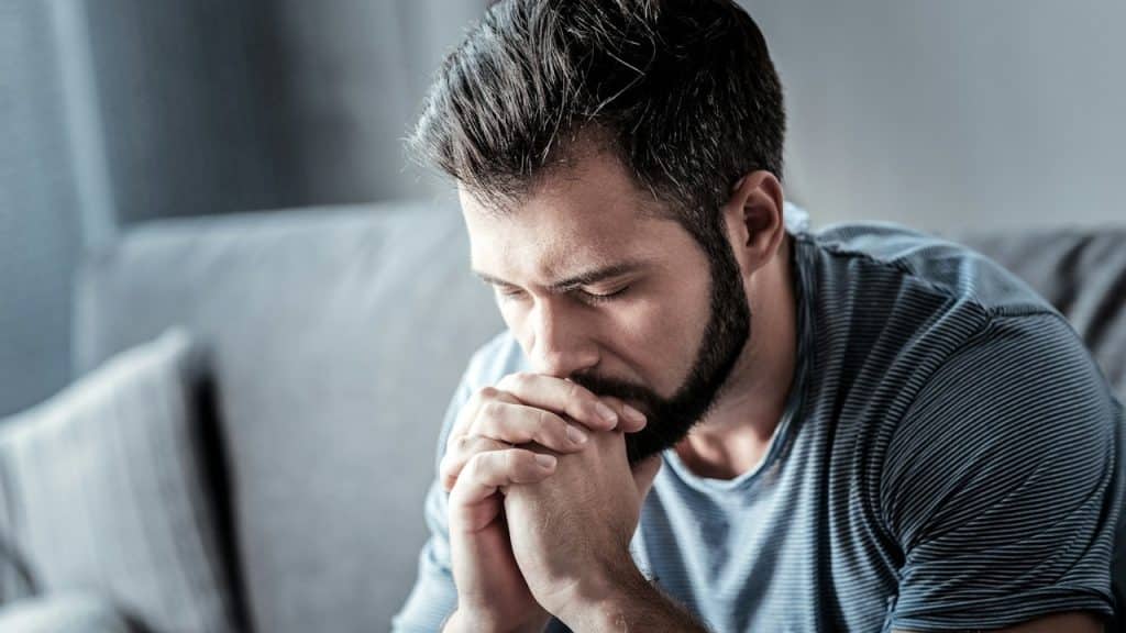 A bearded man looking distressed while sitting on a couch.