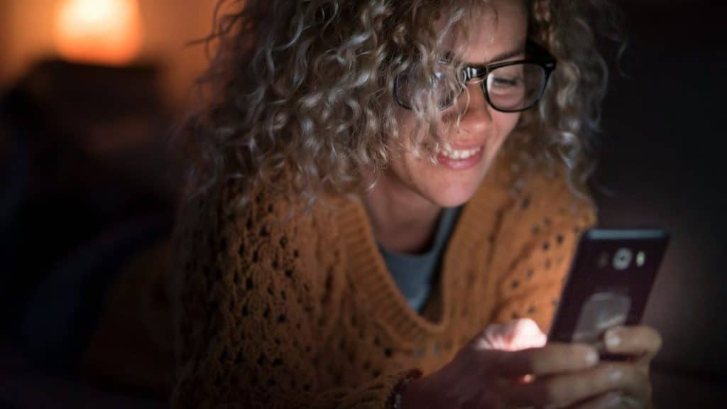 Woman with curly hair and glasses smiling while looking at a phone screen in the dark.