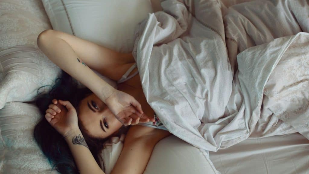 Woman with dark hair lying in bed under white covers, covering part of her face.