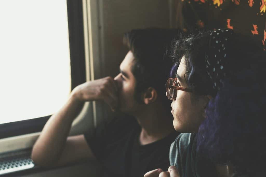 A man gazing out of a window while a woman with glasses looks at him.