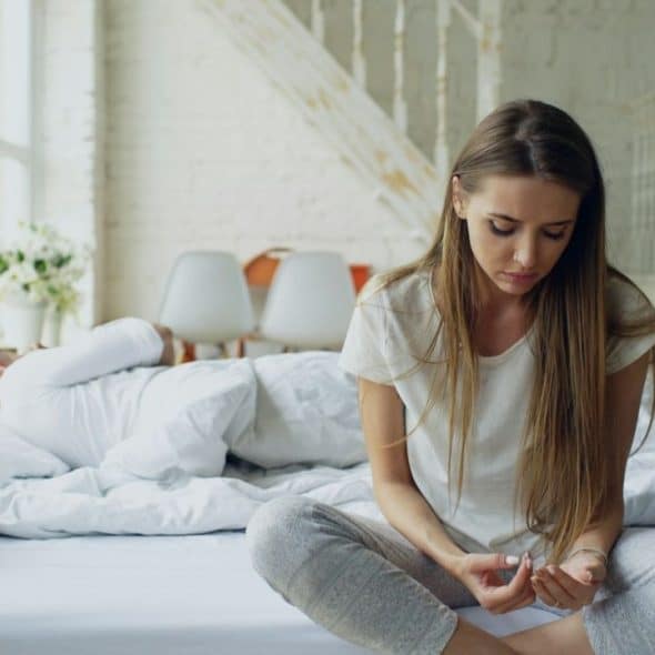 Woman sitting cross-legged on a bed looking down, with a man sleeping behind her.