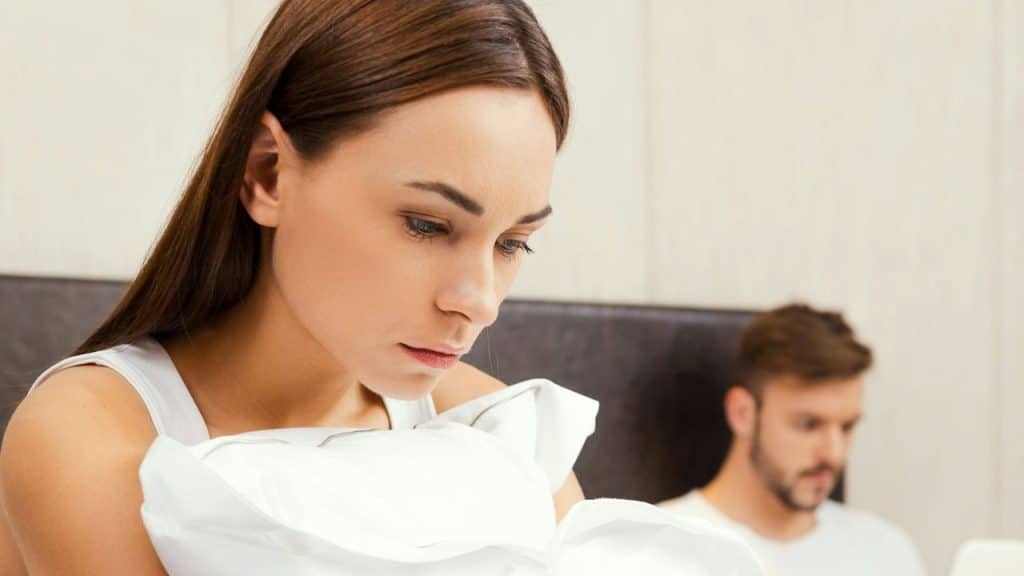 A distressed woman holds a pillow in the foreground, with a man out of focus in the background.