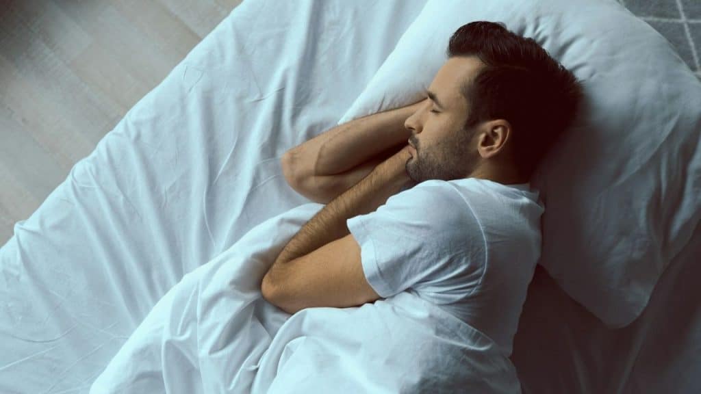 A man with a beard is sleeping on his side in a bed with white sheets and pillows.