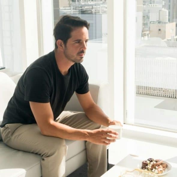 A man sits on a white couch, holding a cup, looking out a large window.