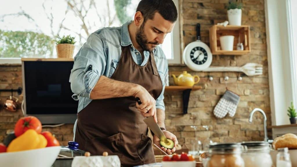 A man in a denim shirt and apron slicing an avocado in a rustic kitchen.