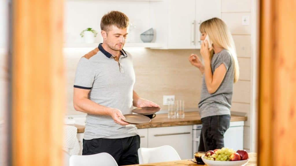 A man sets plates at a table while a woman talks on a phone in the background.