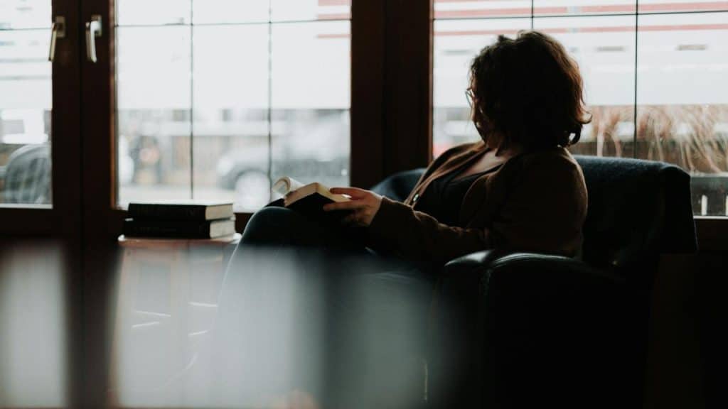 A woman sitting by a window in an armchair while reading a book.