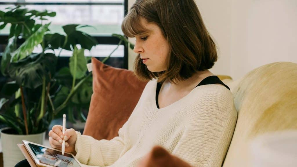 A woman sitting on a couch drawing on a tablet with a stylus.