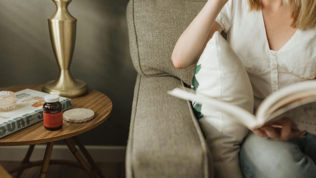 A woman sitting on a couch reading a book beside a small side table.