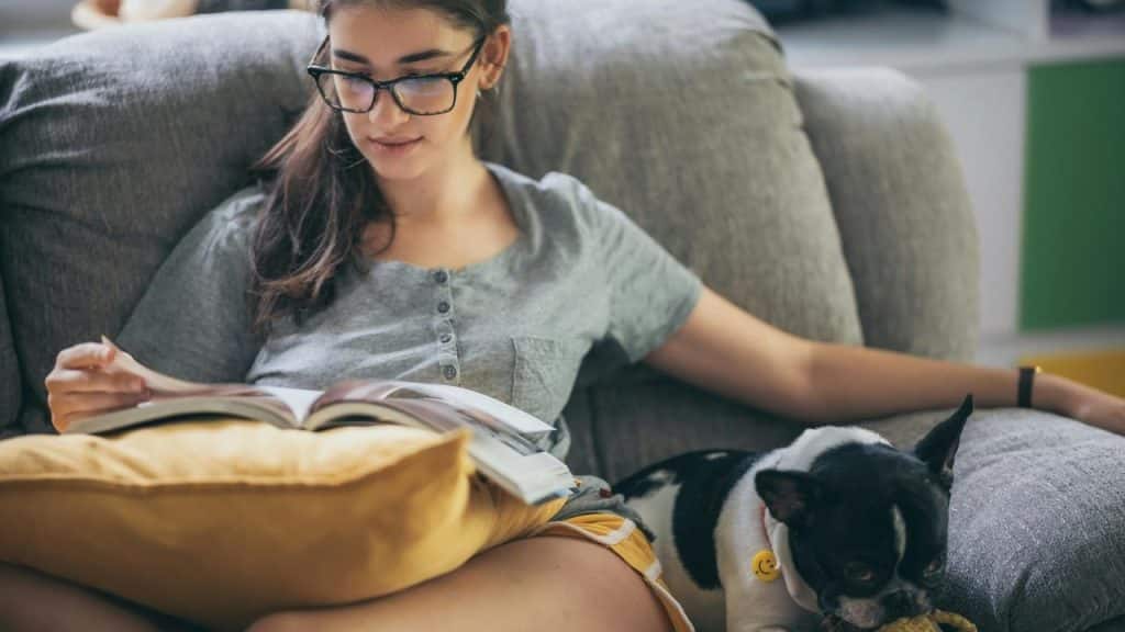 A woman reading a book on a couch with a dog beside her.
