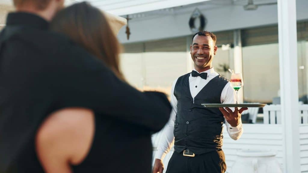 A smiling waiter serving a couple with drinks outside.