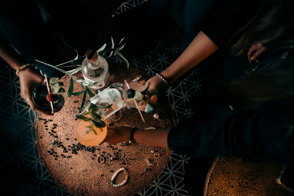 An overhead view of three people's hands reaching toward a round, coppery table with cocktails and a bottle of spirits.