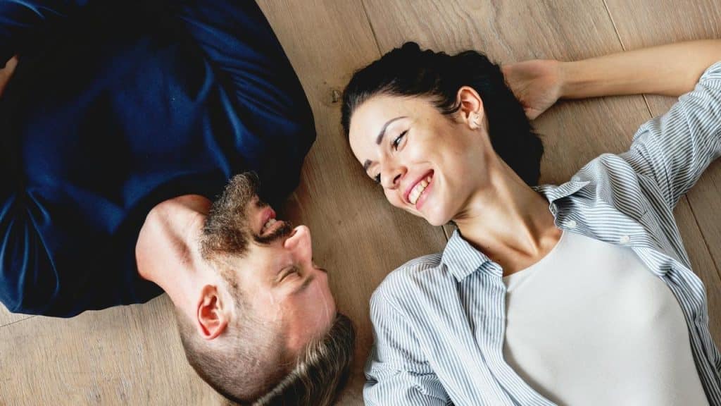 A man and woman lying on a wooden floor, happily staring at each other.