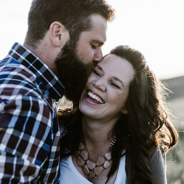 A bearded man kisses a laughing woman on the forehead in a sunny outdoor field.