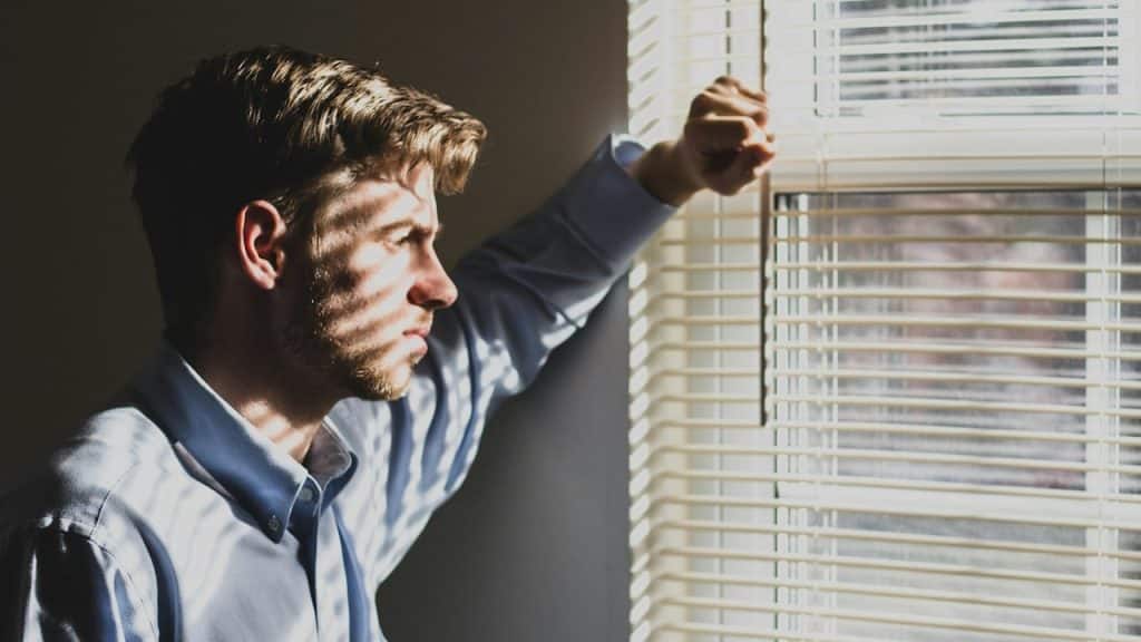 A man looking out of a window that has blinds on it.
