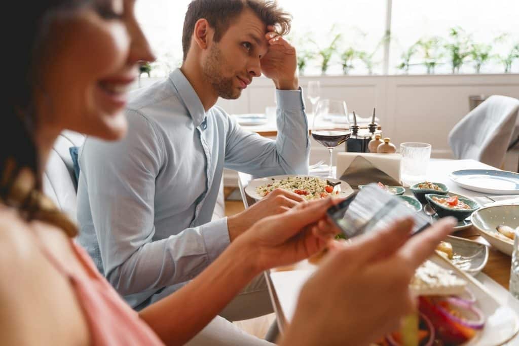 The man looks annoyed while the woman is taking photos of the food.