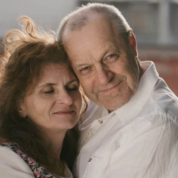 A close-up of a smiling, older, fair-skinned couple embracing outdoors.