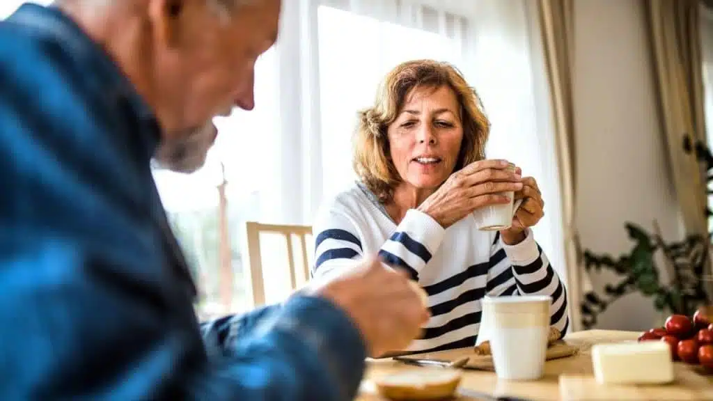 Mature couple at a table, woman drinking coffee, man eating, natural light.