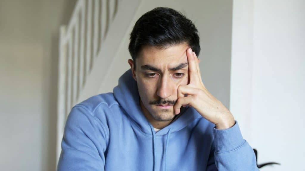 Man in a blue hoodie resting his forehead on his hand, looking down thoughtfully.