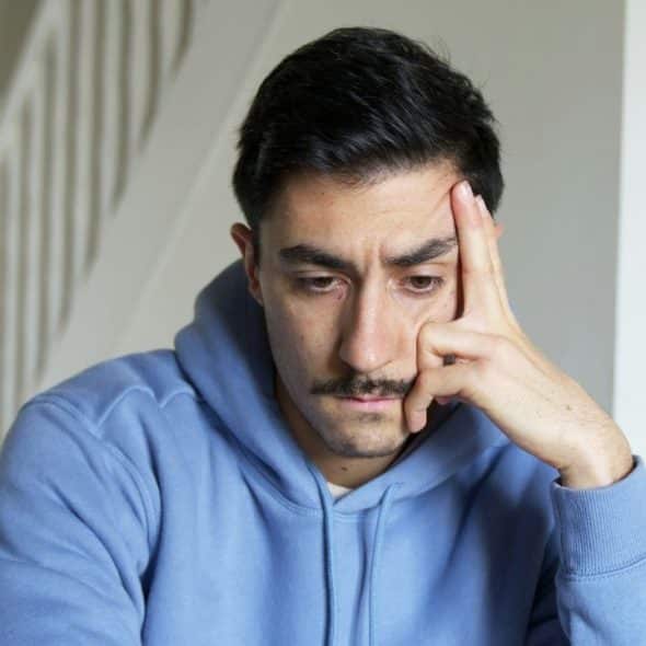 Man in a blue hoodie resting his forehead on his hand, looking down thoughtfully.