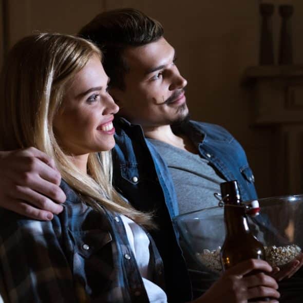 Couple watching TV with snacks and drinks.
