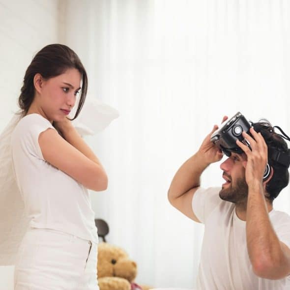 A woman in bed is about to start a pillow fight with her husband.