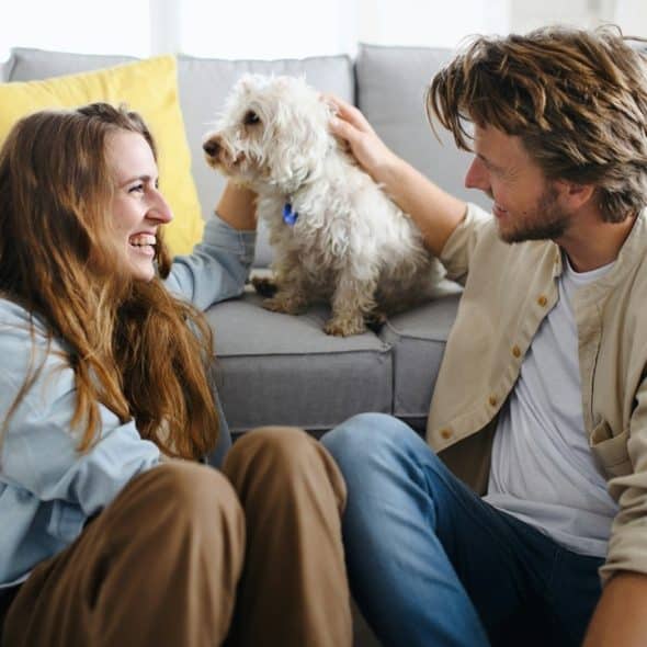 A couple smiling and sitting on the floor while the man strokes a dog who’s sitting on a couch.