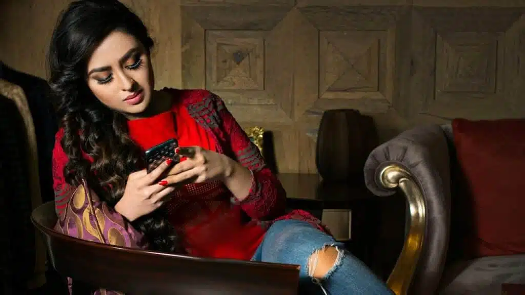 A woman in a red shirt texting on a phone sitting on a brown wooden armchair.