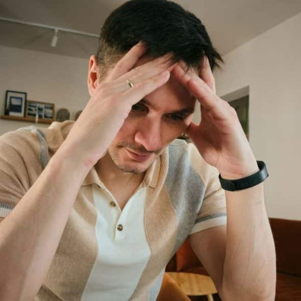 Man indoors, holding his head with both hands, looking down with a pained expression.