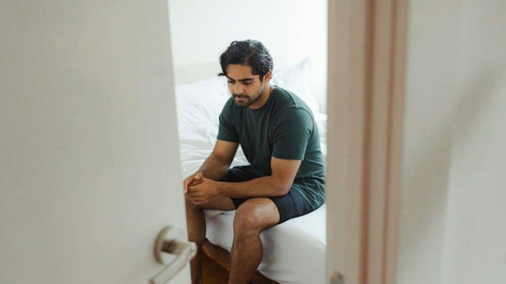 Man in a dark shirt and shorts sitting alone on a bed, viewed through an open doorway.