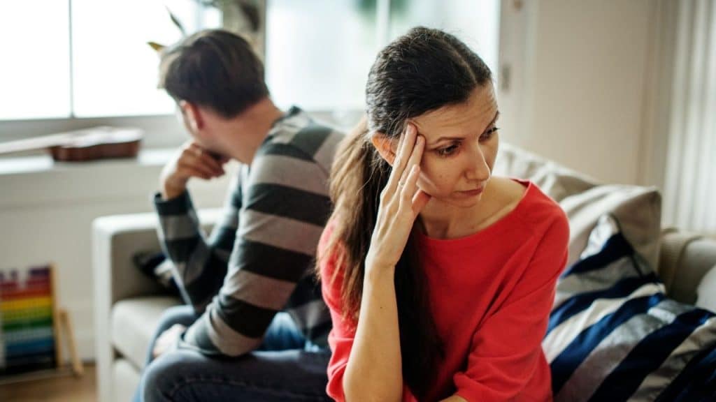 A woman in a red shirt with a hand on her temple sits back-to-back with a man.