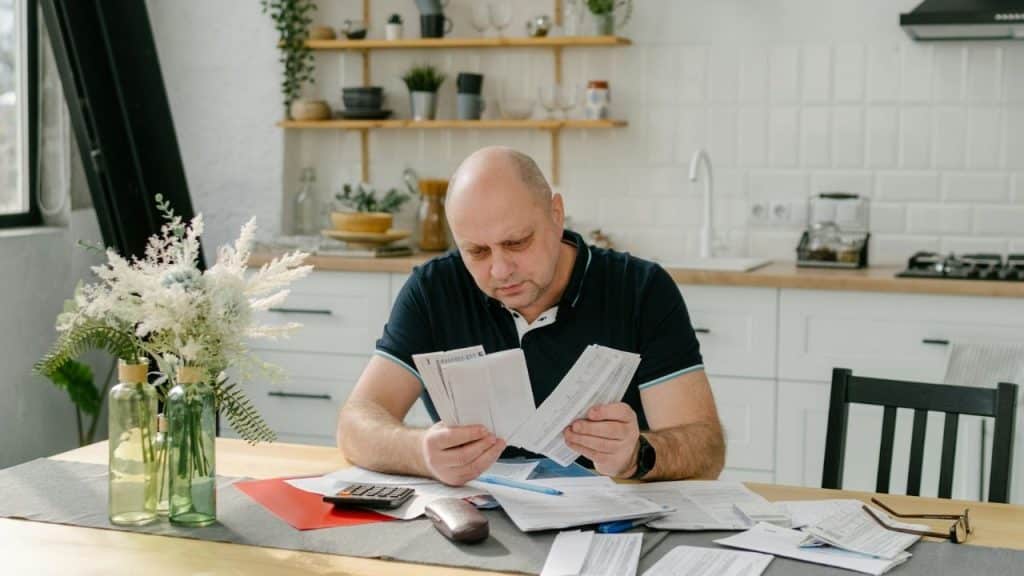 Middle-aged bald man sitting at a kitchen table, looking intensely at several bills and papers.