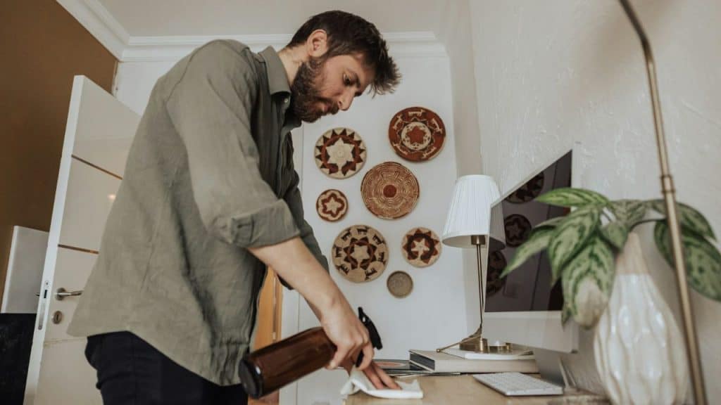 A man in a green shirt sprays and wipes a wooden desk with a white cloth.