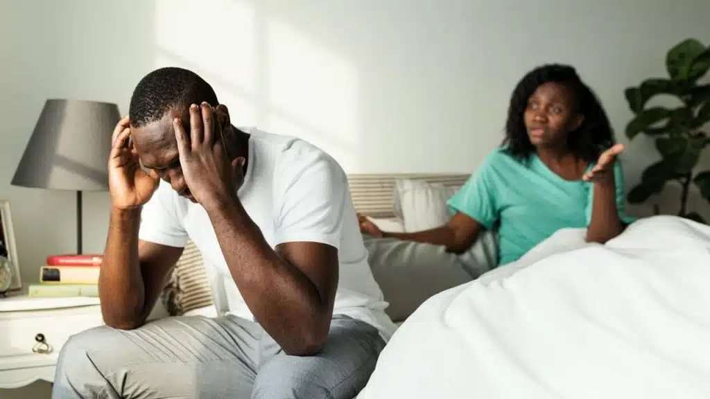 A man sits on the edge of a bed, holding his head, while a woman argues behind him.