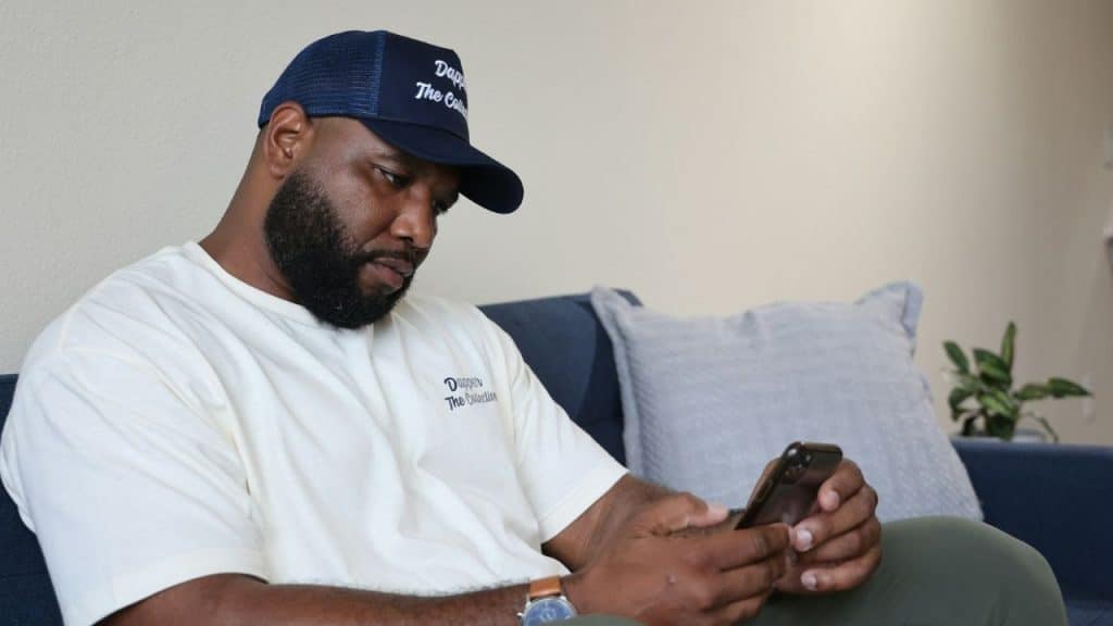 Bearded man wearing a trucker hat and white shirt, looking down at his phone indoors.