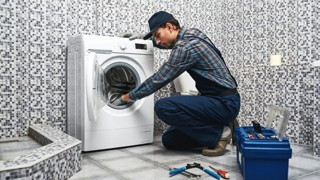 A man kneels in a tiled bathroom, working on a front-loading washing machine.