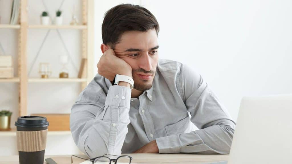 Man looking wearily at a laptop screen, resting his head on his hand.