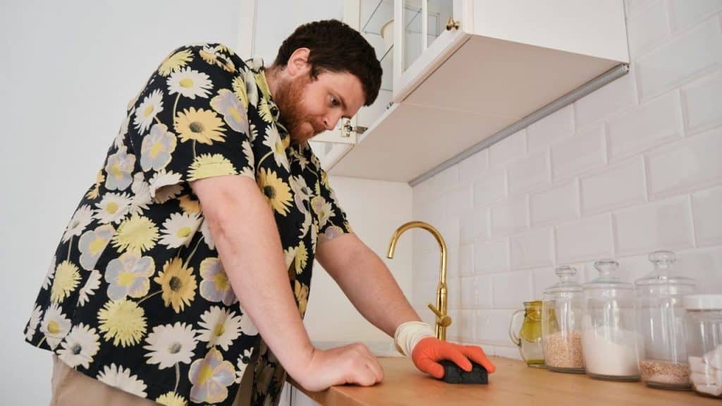 Man in a floral shirt and rubber glove scrubbing a wooden kitchen countertop.