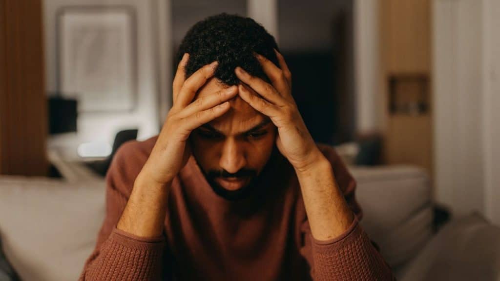 Man sitting indoors in dim lighting, hands pressing against his head in distress.
