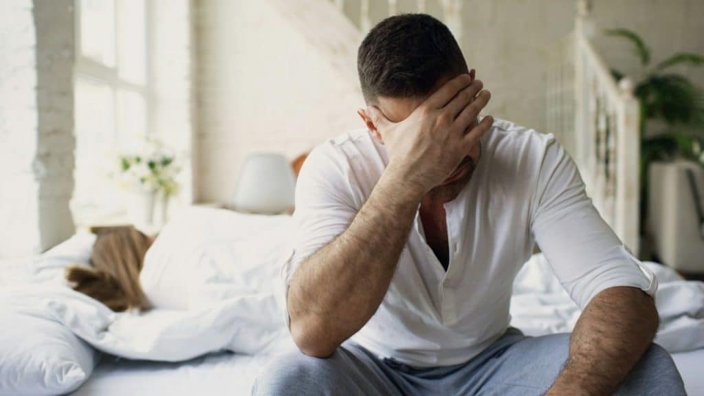Man sitting on a bed covering his face with his hand while a person sleeps behind him.