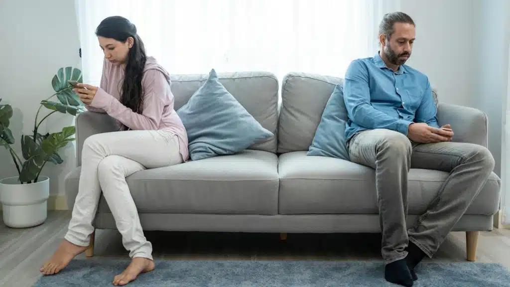 Couple sitting far apart on a couch, both looking at their phones, avoiding eye contact.