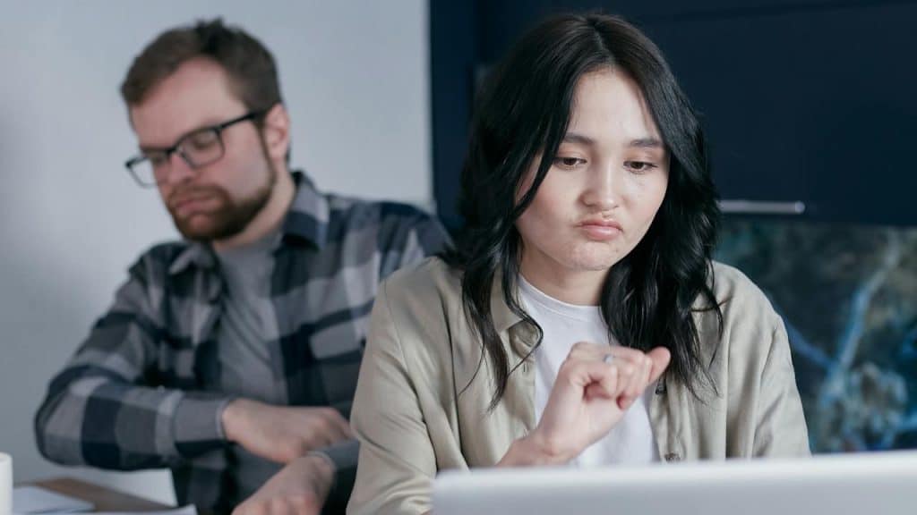 Woman looking disgruntled while working on a laptop, with a man looking at papers in the background.