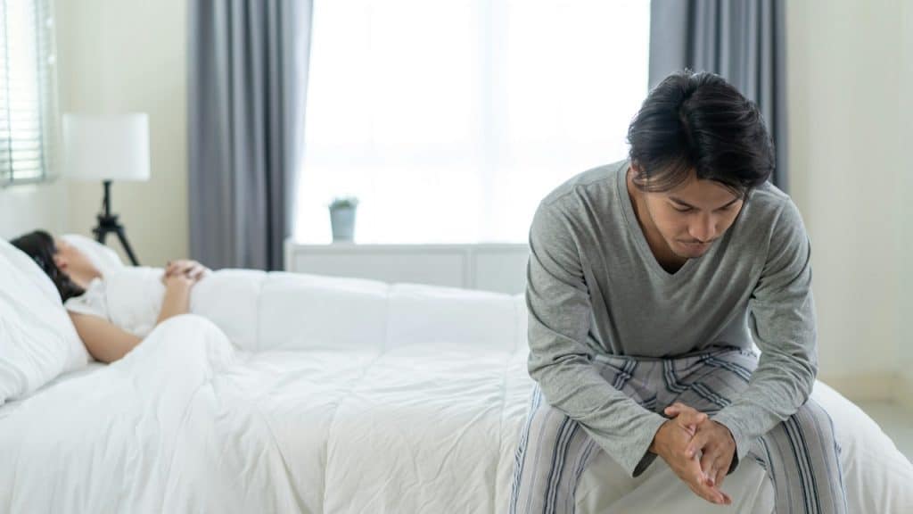 Man sitting sadly on the edge of a bed while a woman sleeps in the background.