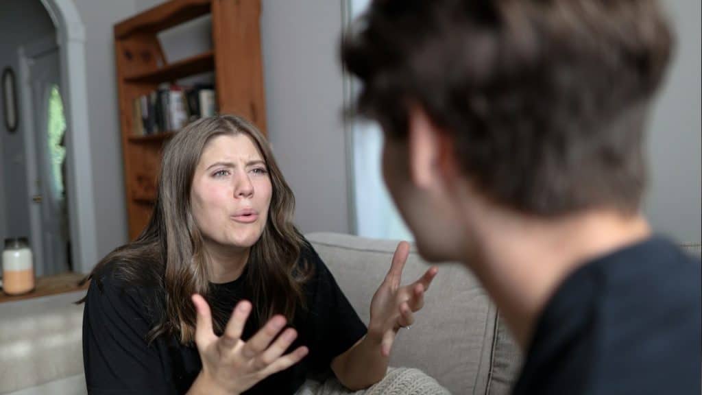 Woman sitting on a couch, passionately arguing and gesturing with her hands towards a blurred man.